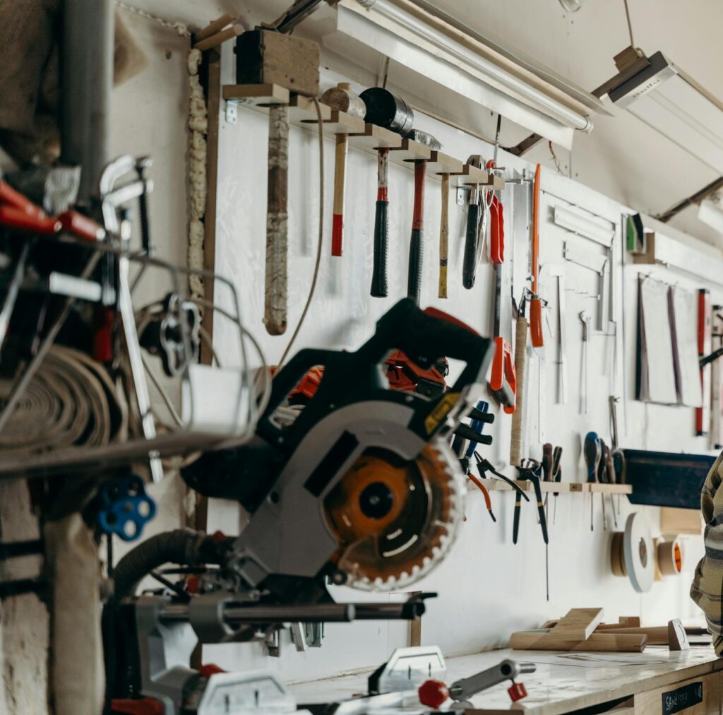 A man with long hair and beanie stands in a workshop with tools on the wall, reflecting a warm workspace atmosphere.